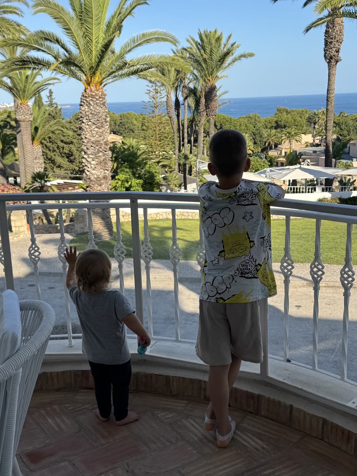 Zwei Kinder blicken von einer Terrasse über Palmen auf das Meer